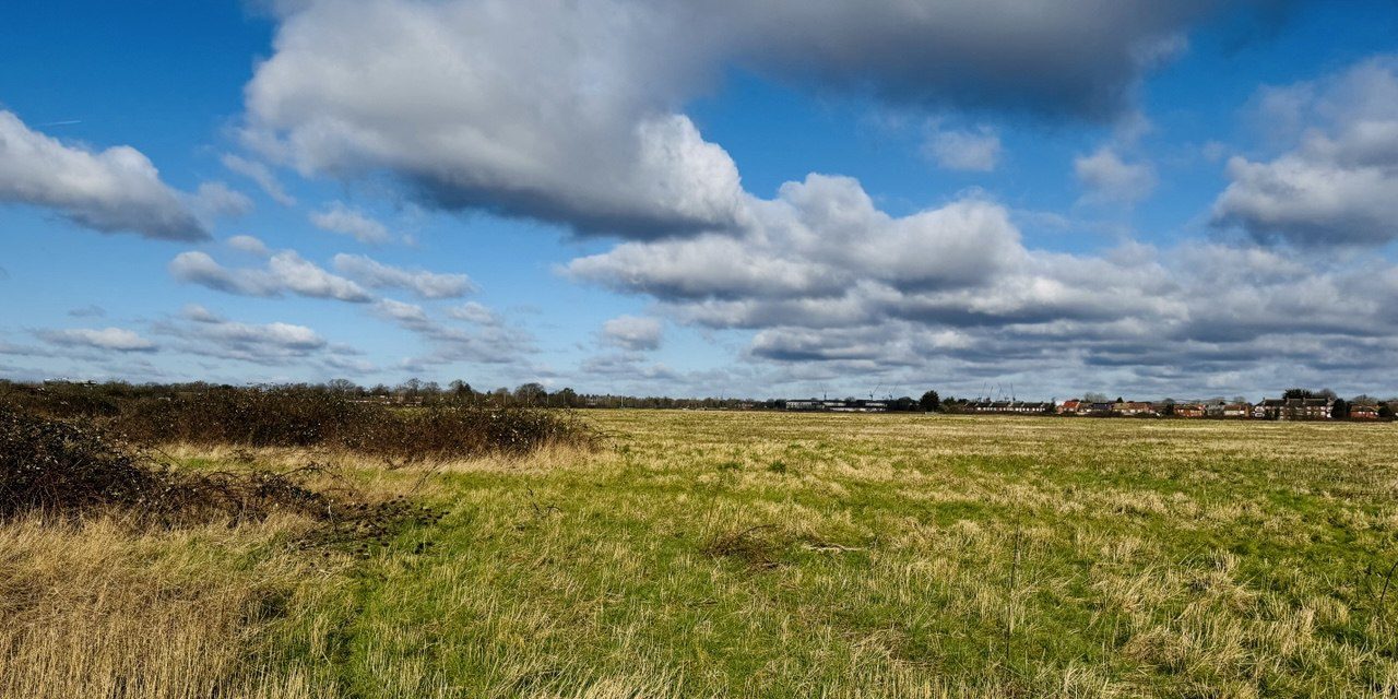 photo_2025-04-02 13.14.36 Expansive open field with tall grass under a partly cloudy sky, with distant houses on the horizon.